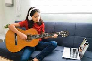 Fille latine avec des écouteurs écoutant ses cours de musique en ligne. Enfant artistique jouant de la guitare acoustique et apprenant les accords