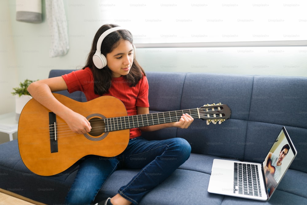 Lateinamerikanisches Mädchen mit Kopfhörern, das ihren Online-Musikunterricht hört. Künstlerisches Kind, das akustische Gitarre spielt und die Akkorde lernt