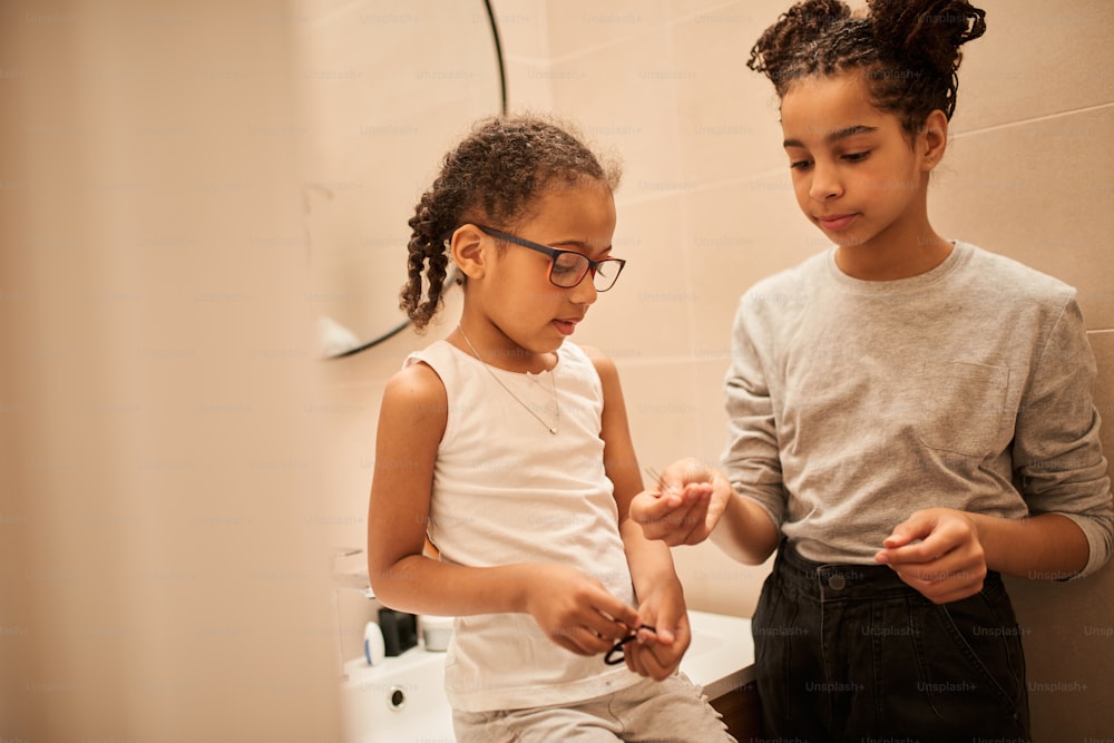 My lovely sister. Waist up portrait view of the little multiracial girl wearing glasses sitting at the bathroom and waiting while her sister making braids for her. Stock photo