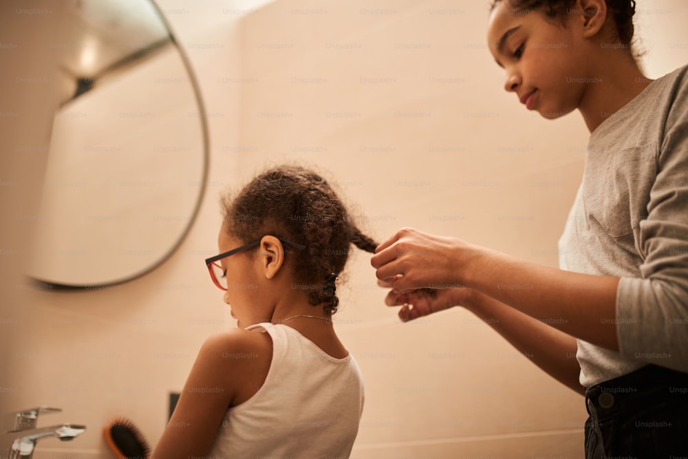 Sister support. Low angle view of the multiracial girl combing hair of her little sister at the bathroom and weaving braids for she before the walking. Stock photo
