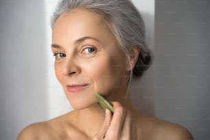 Vista verticale di una donna sorridente dai capelli grigi maturi che utilizza un massaggiatore viso al quarzo per il trattamento della pelle mentre guarda la fotocamera nello studio dietro il muro bianco. Immagine