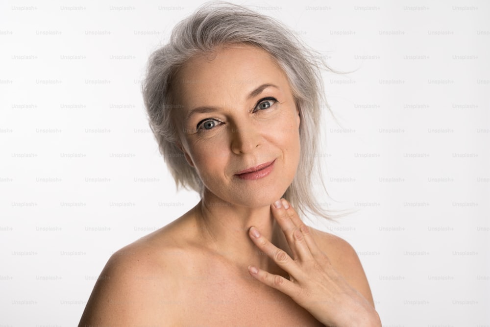 Isolated shot of cheerful aged woman with healthy glowing skin and happy smile touching neck gently at the studio with white background. Beautiful european female has well groomed complexion and body