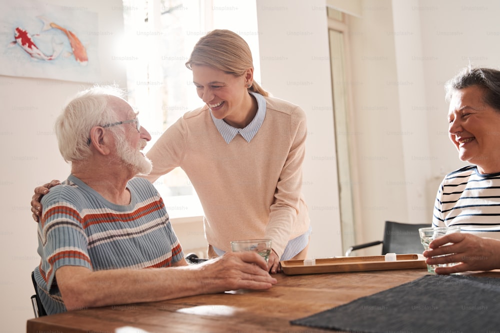 All will be ok. Waist up portrait view of the happy senior man looking at the nurse embracing him while sitting at the table with his female friend. Stock photo