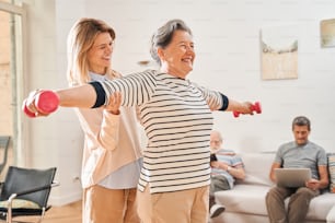 With dumbbells. Happy mature woman training with dumbbells with her caregiver at the nurse house. She including pensioners in activity. Domestic life in retirement house concept