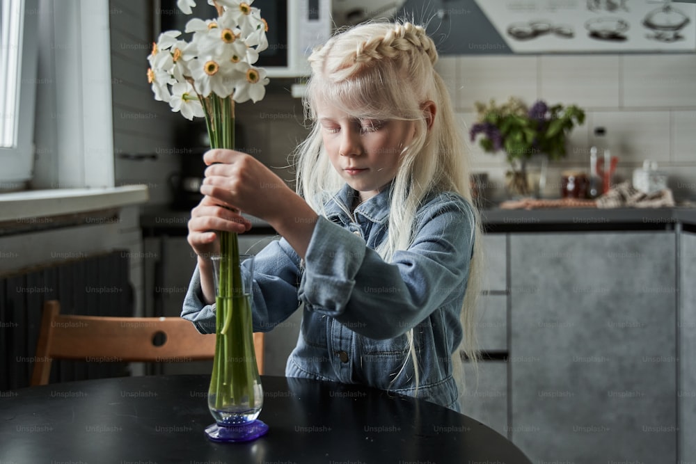 Doing diligent. Waist up portrait view of the calm blonde girl standing at the table and putting flowers at the vase while helping to her father at the kitchen. Floristic and home concept