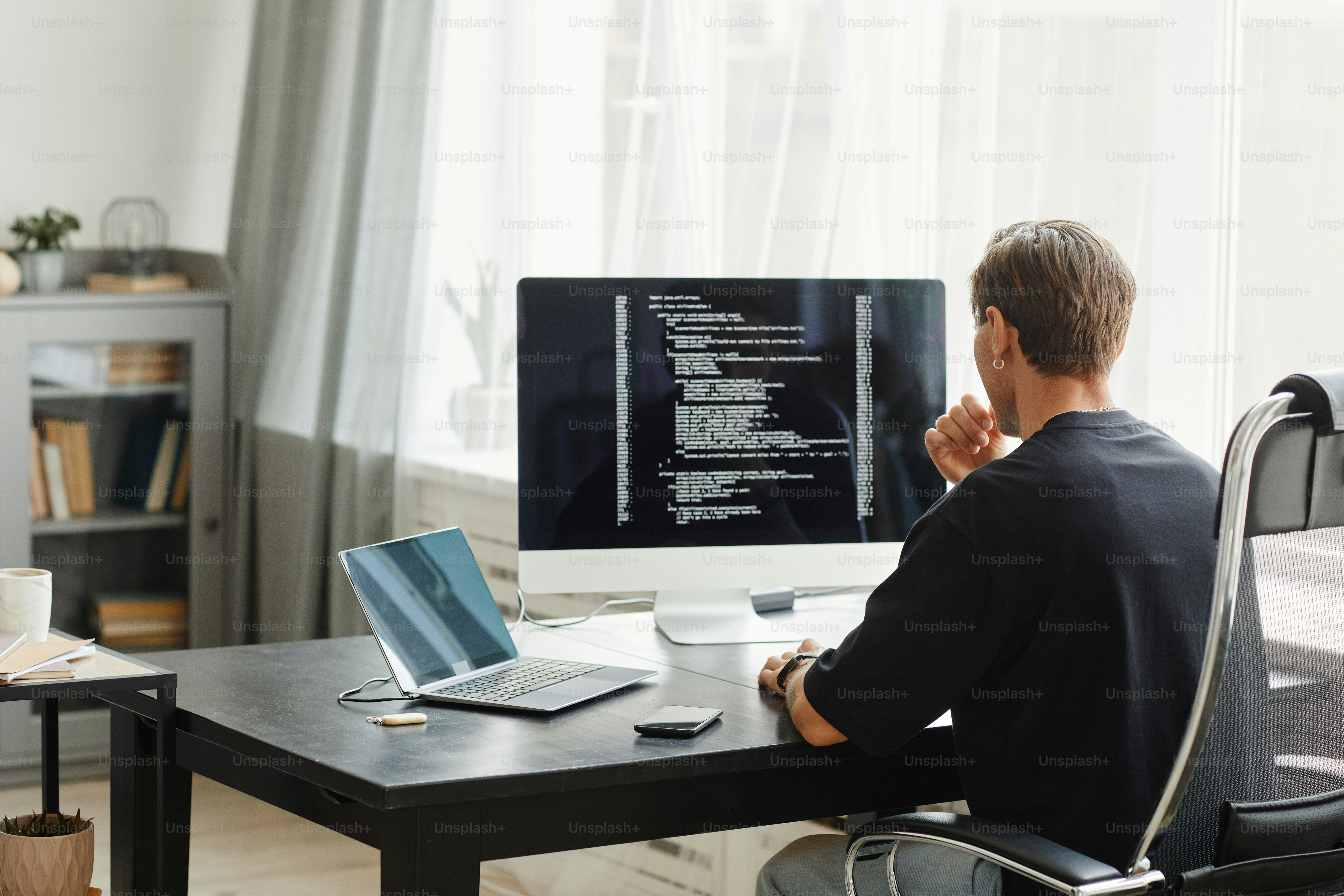 Rear view of computer programmer sitting at the table in front of computer monitor and developing software at office