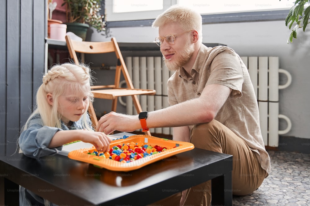 Kindheit. Ganzkörperansicht des kleinen Albino-Kindes, das Details ihres Spiels faltet und am Tisch sitzt, während ihr blonder Vater ihr mit Lustgefühlen hilft. Familien- und Freizeitspielkonzept