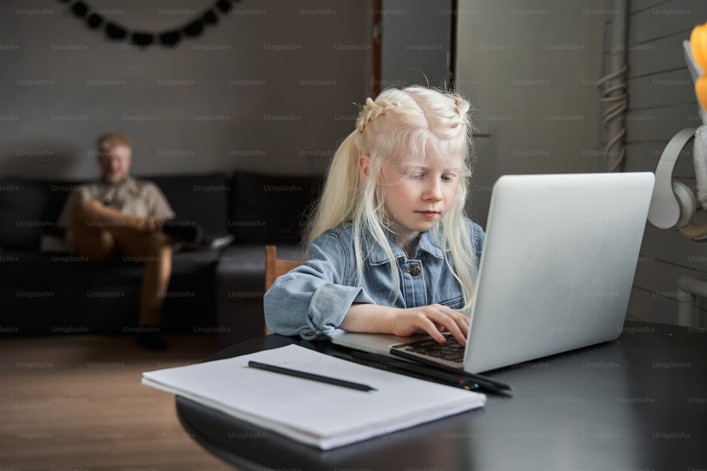 Thoughtful little albino caucasian girl stuck with hard task, preparing homework at home at the self isolation while her father relaxing at the background. Education and school concept