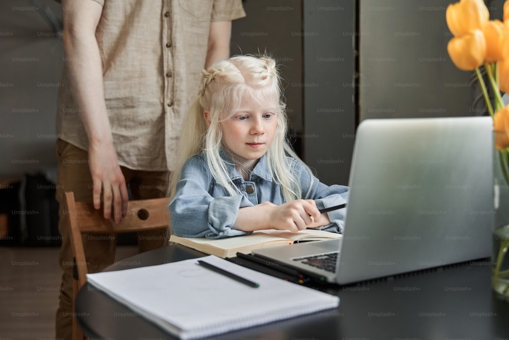 Petite fille albinos caucasienne souriante ayant une éducation à distance avec professeur utilisant un ordinateur portable. Happy petit enfant étudier en ligne sur ordinateur avec son père en arrière-plan. Concept d’enseignement à domicile