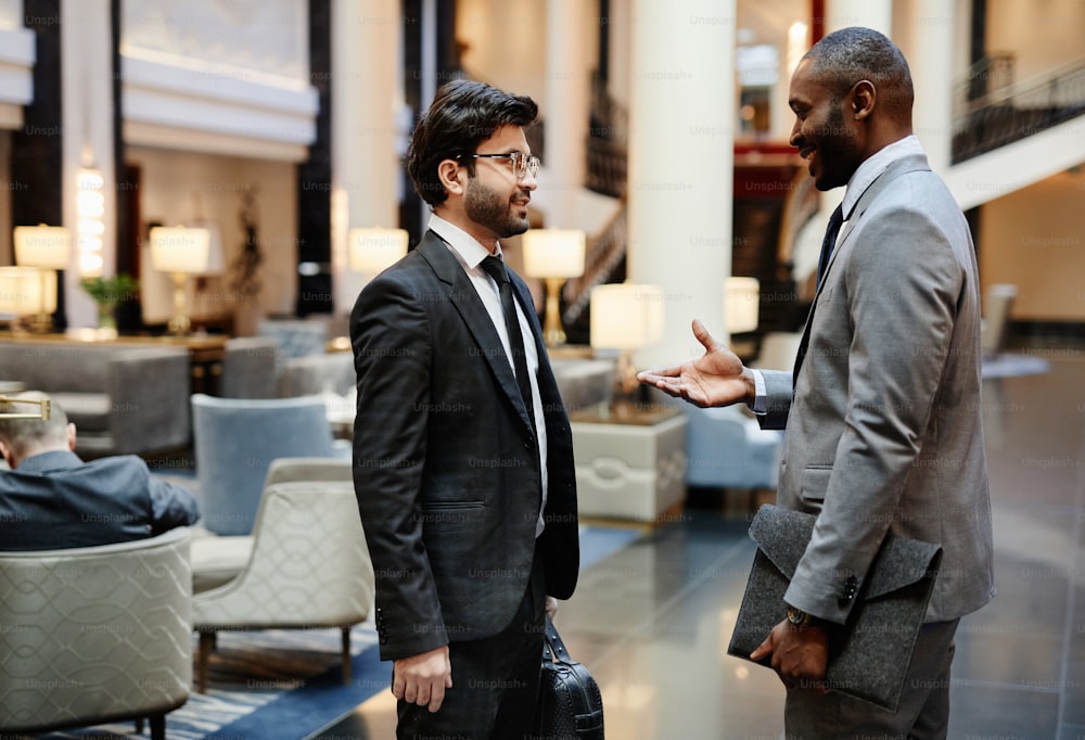 Side view portrait of two successful businessmen discussing work while standing in hotel lobby, copy space