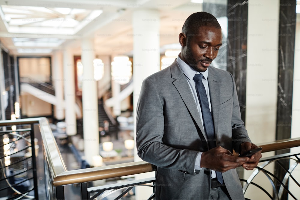 Portrait à la taille d’un homme d’affaires afro-américain debout au balcon et utilisant un smartphone dans le hall luxueux de l’hôtel, espace de copie