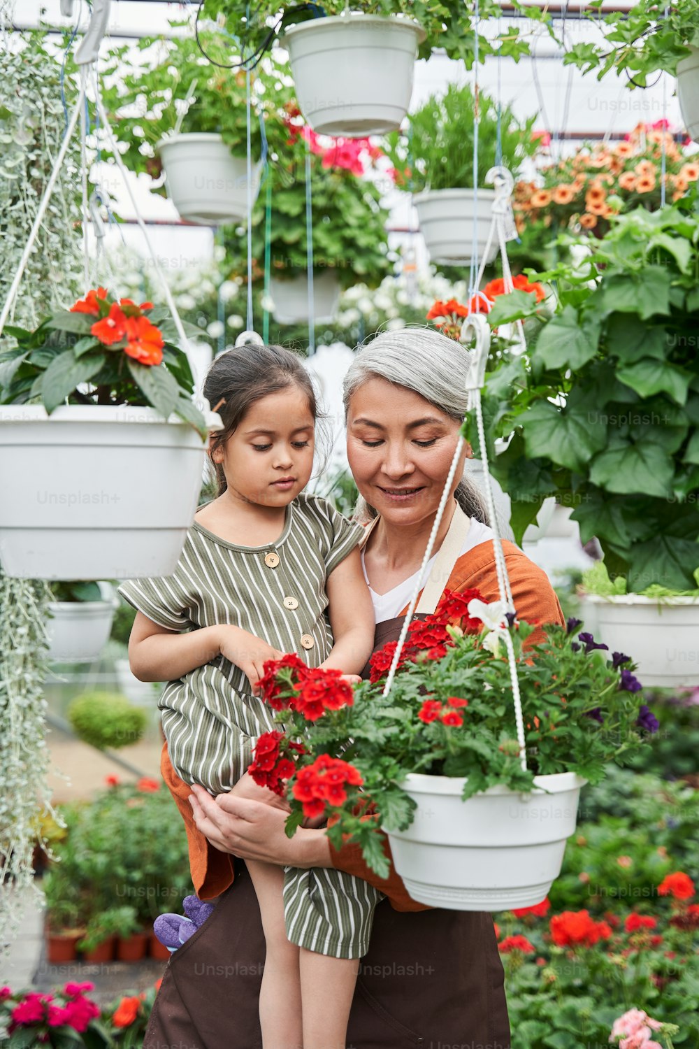 Waist up portrait view of the senior lady embracing her little granddaughter and telling something to her while spending time at the greenhouse. Stock photo