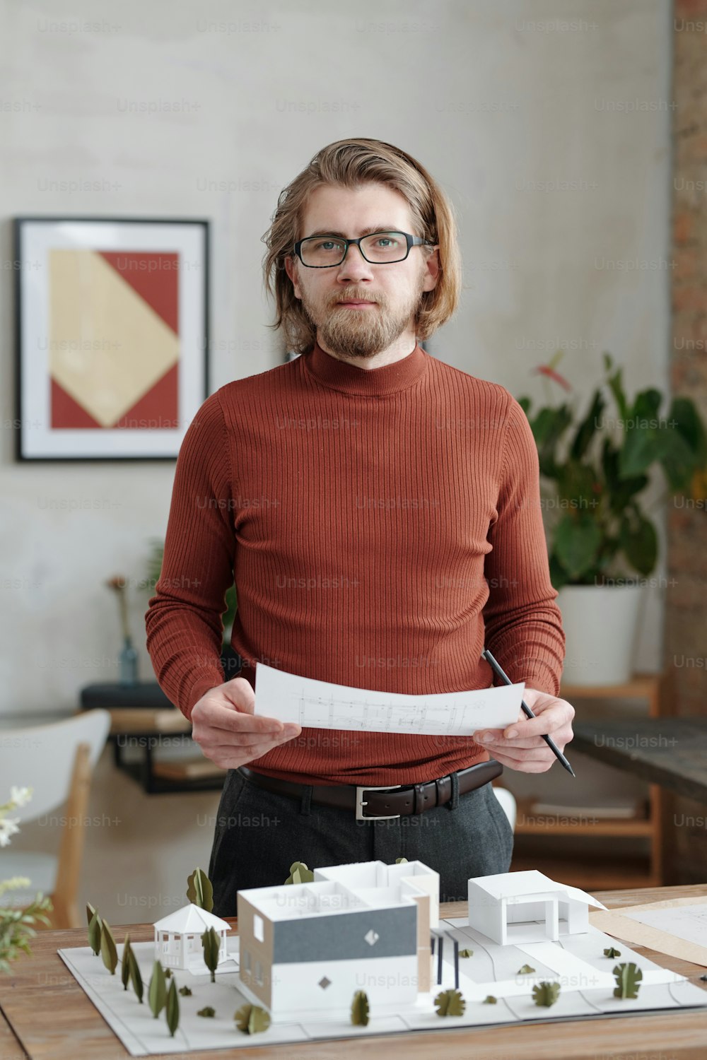 Portrait of young Caucasian creative architect in turtleneck standing at desk and comparing sketch and maquette