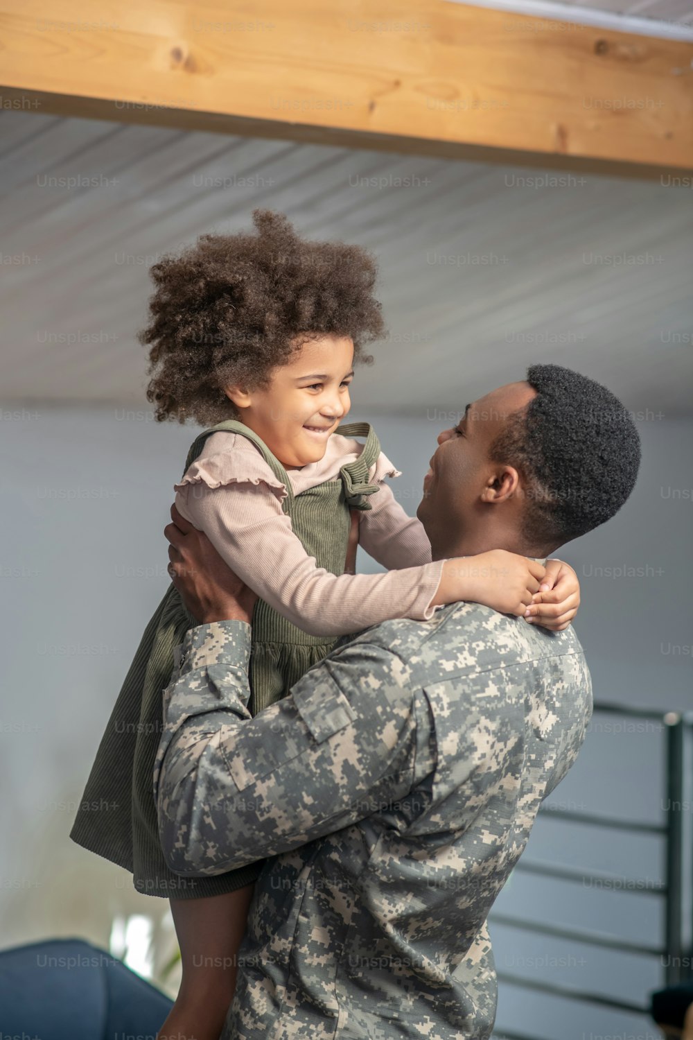 Reunião muito esperada. Jovem pai forte de pele escura em uniforme militar segurando a pequena filha de cabelos cacheados brilhantes regozijando-se com o encontro