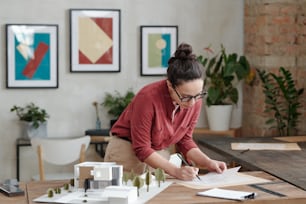 Busy young female architect with hair bun standing at table with 3D model of cottage house and editing building plan