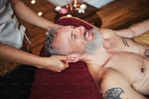 Vista superior do homem bonito barbudo sorridente deitado no travesseiro enquanto o massagista asiático está tratando suas orelhas