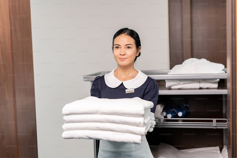 Bonito personal de servicio de habitaciones sosteniendo sábanas limpias mientras está de pie junto a la puerta abierta