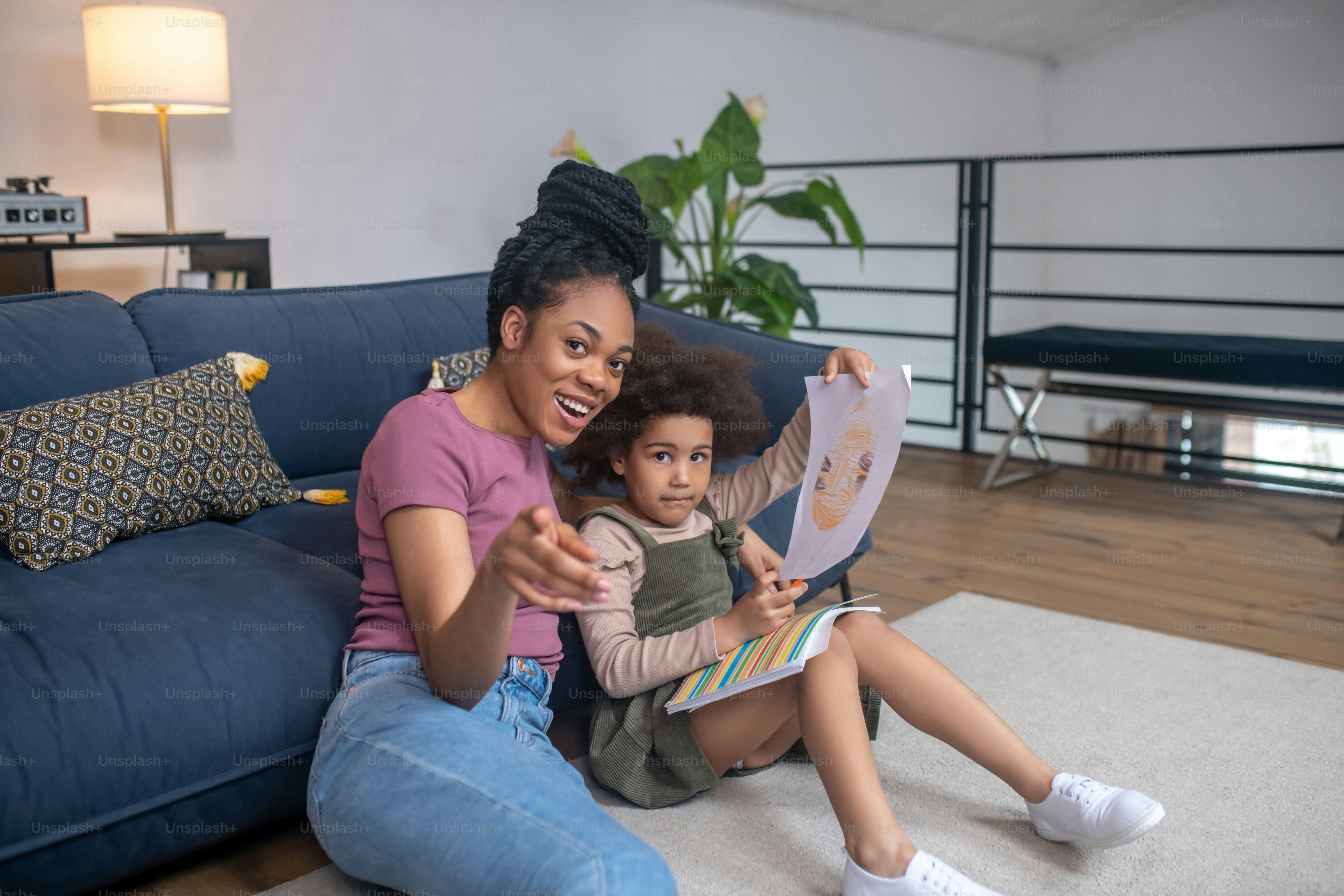 Attention. Joyful dark-skinned young woman pointing forward sitting on floor with her little daughter looking intently and holding picture