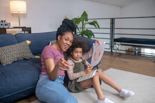 Attention. Joyful dark-skinned young woman pointing forward sitting on floor with her little daughter looking intently and holding picture