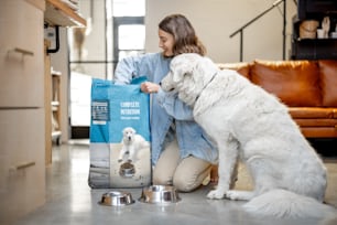 Una joven alimenta a su enorme perro blanco con un alimento seco, sentado en el suelo con un paquete de comida para mascotas en casa. Concepto de nutrición sana y equilibrada para mascotas