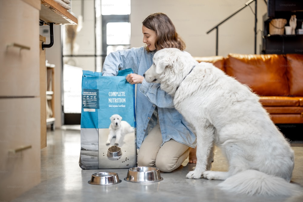 Young woman feeds her huge white dog with a dry food, sitting on the floor with a package of pet's food at home. Concept of healthy and balanced nutrition for pets