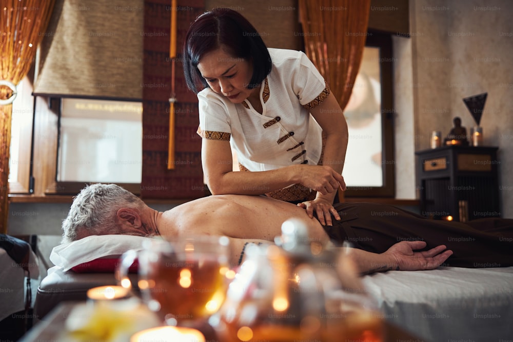 Une jolie femme asiatique fait un massage thaïlandais avec les coudes pour un gars grisonnant tatoué allongé sur le canapé