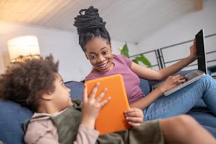 Interesting here. Little curly dark skinned girl showing tablet screen to joyful mom sitting next to laptop on sofa at home