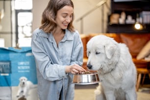 Young woman feeds her huge white dog with a dry food, sitting on the floor with a package of pet's food at home. Concept of healthy and balanced nutrition for pets