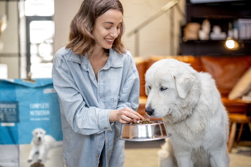 Young woman feeds her huge white dog with a dry food, sitting on the floor with a package of pet's food at home. Concept of healthy and balanced nutrition for pets