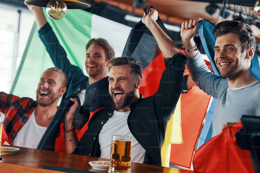 Jubelnde junge Männer, die in internationale Flaggen gehüllt sind und Bier genießen, während sie in der Kneipe Sportspiele verfolgen