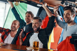 Giovani uomini chemasi coperti di bandiere internazionali che si godono la birra mentre guardano la partita sportiva nel pub