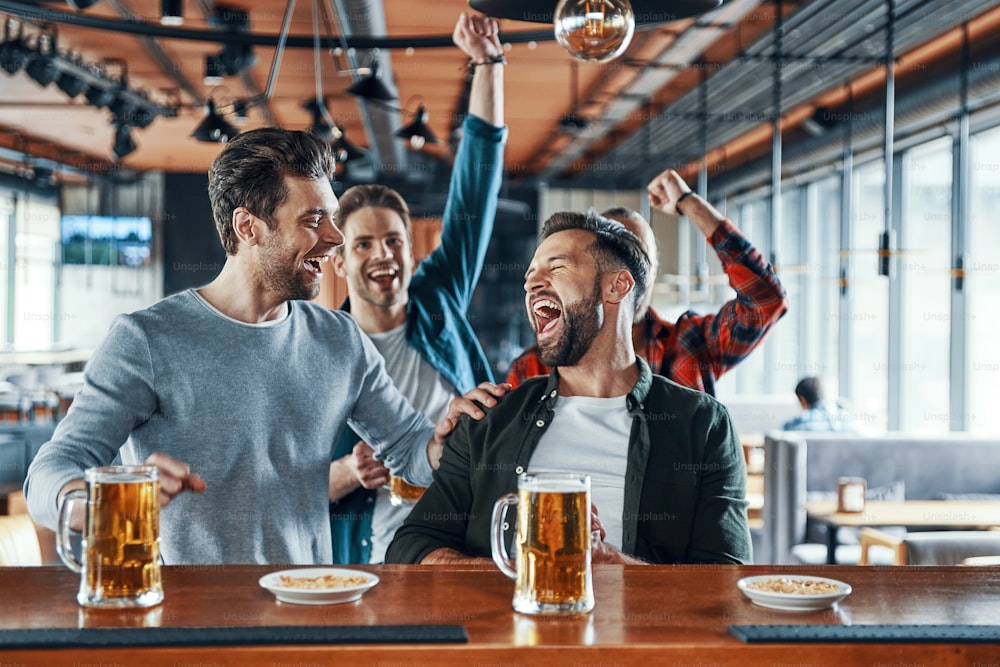 Excited young men in casual clothing drinking beer and watching sport game while sitting in the pub