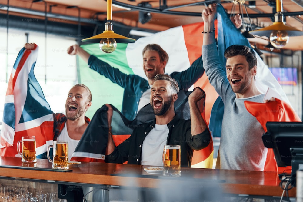 Jubelnde junge Männer, die in internationale Flaggen gehüllt sind und Bier genießen, während sie in der Kneipe Sportspiele verfolgen