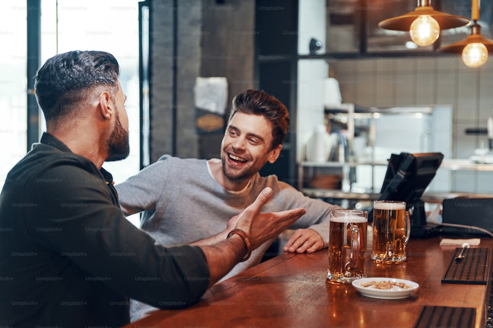 Two happy young men in casual clothing talking and drinking beer while spending time in the pub