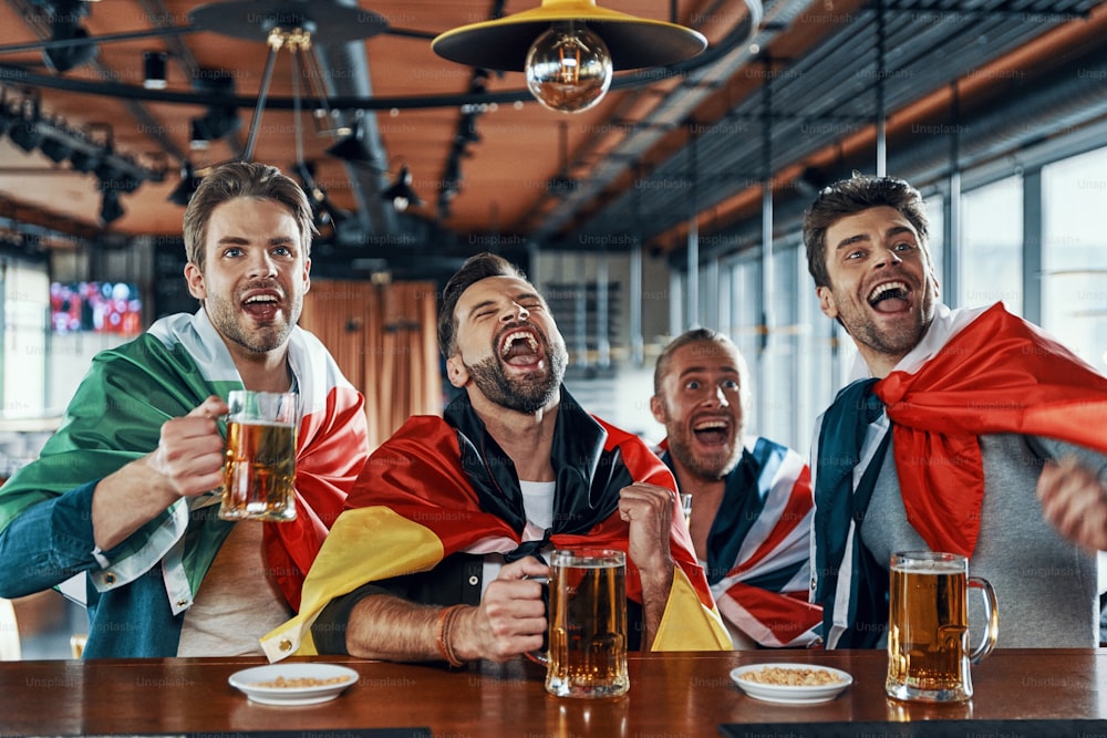 Des jeunes hommes heureux couverts de drapeaux internationaux buvant de la bière et regardant un match de sport assis dans le pub