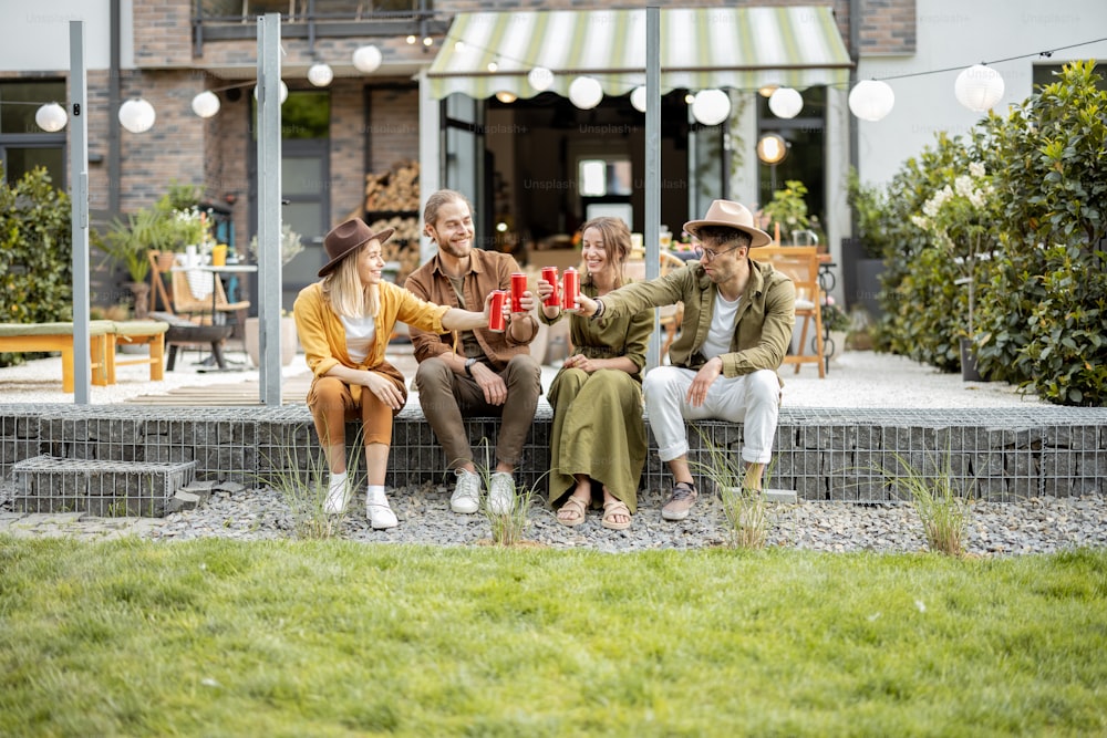 Grupo de jóvenes amigos que pasan el verano juntos, divirtiéndose mientras están sentados en una fila con bebidas en un porche de la casa de campo