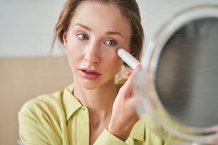 Porträtansicht einer brünetten, aufmerksamen Frau mit Vitiligo-Muttermalen, die den Spiegel hält und hineinschaut, während sie drinnen eine Gesichtsmassage macht. Konzept von Schönheit, Selbstpflege, Kosmetik