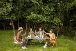 Gruppo di giovani che fanno una cena festosa nella natura, seduti insieme a un tavolo su un prato verde vicino alla foresta. Ampia visualizzazione, spazio di copia