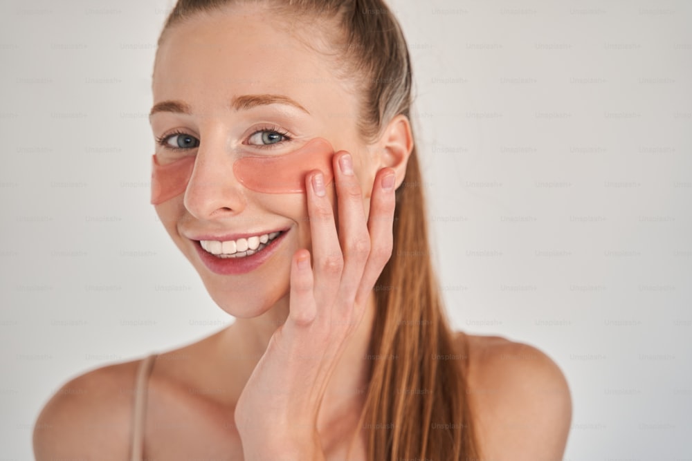 Portrait view of happy young woman wearing under eye patches touching her skin with satisfaction. She is standing with naked shoulders and smiling to the camera