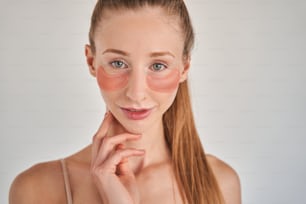 Portrait view of happy young woman wearing under eye patches touching her skin with satisfaction. She is standing with naked shoulders and smiling to the camera