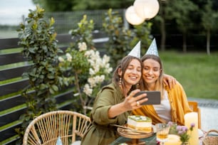 Portrait de deux amies célébrant leur anniversaire, faisant un selfie ou un appel téléphonique assis près d’une table de fête dans la cour arrière à l’extérieur