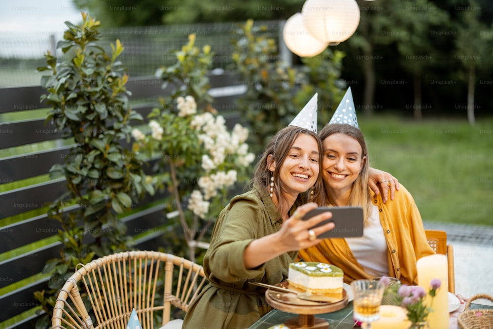 Portrait de deux amies célébrant leur anniversaire, faisant un selfie ou un appel téléphonique assis près d’une table de fête dans la cour arrière à l’extérieur