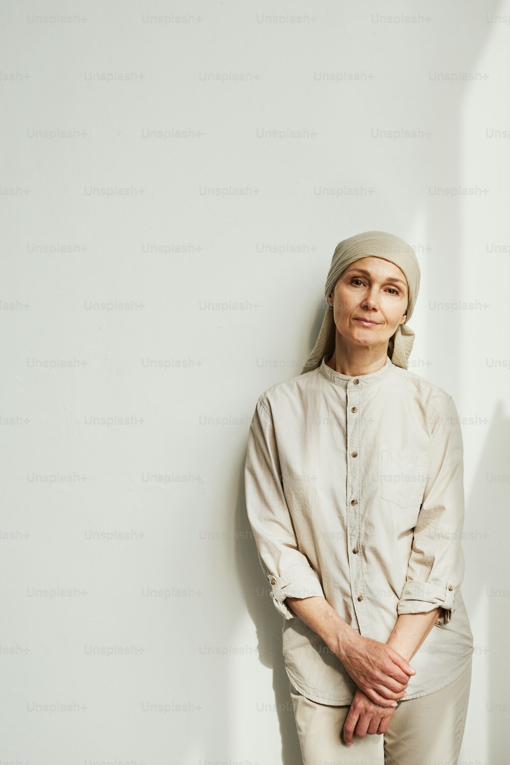 Minimal portrait of pensive mature woman wearing headscarf and looking at camera while standing by white wall in sunlight