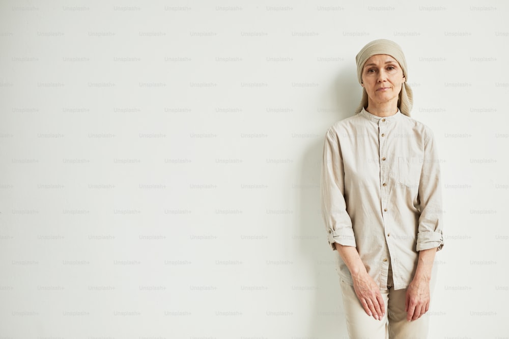 Minimales Porträt einer nachdenklichen reifen Frau, die Kopftuch trägt und in die Kamera schaut, während sie an einer weißen Wand steht, Kopierraum
