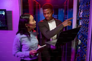 Young Afro-American technician pointing at cables while explaining server productivity to Asian colleague in data center illuminated with neon light