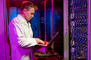 Serious concentrated mature Caucasian IT engineer in white coat standing at cart with server components and using laptop while troubleshooting server problem