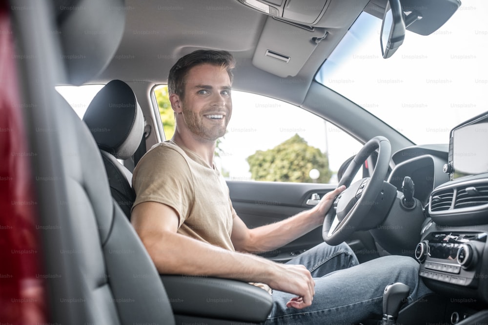 Good mood. Happy attractive young man looking at camera while driving car during daytime