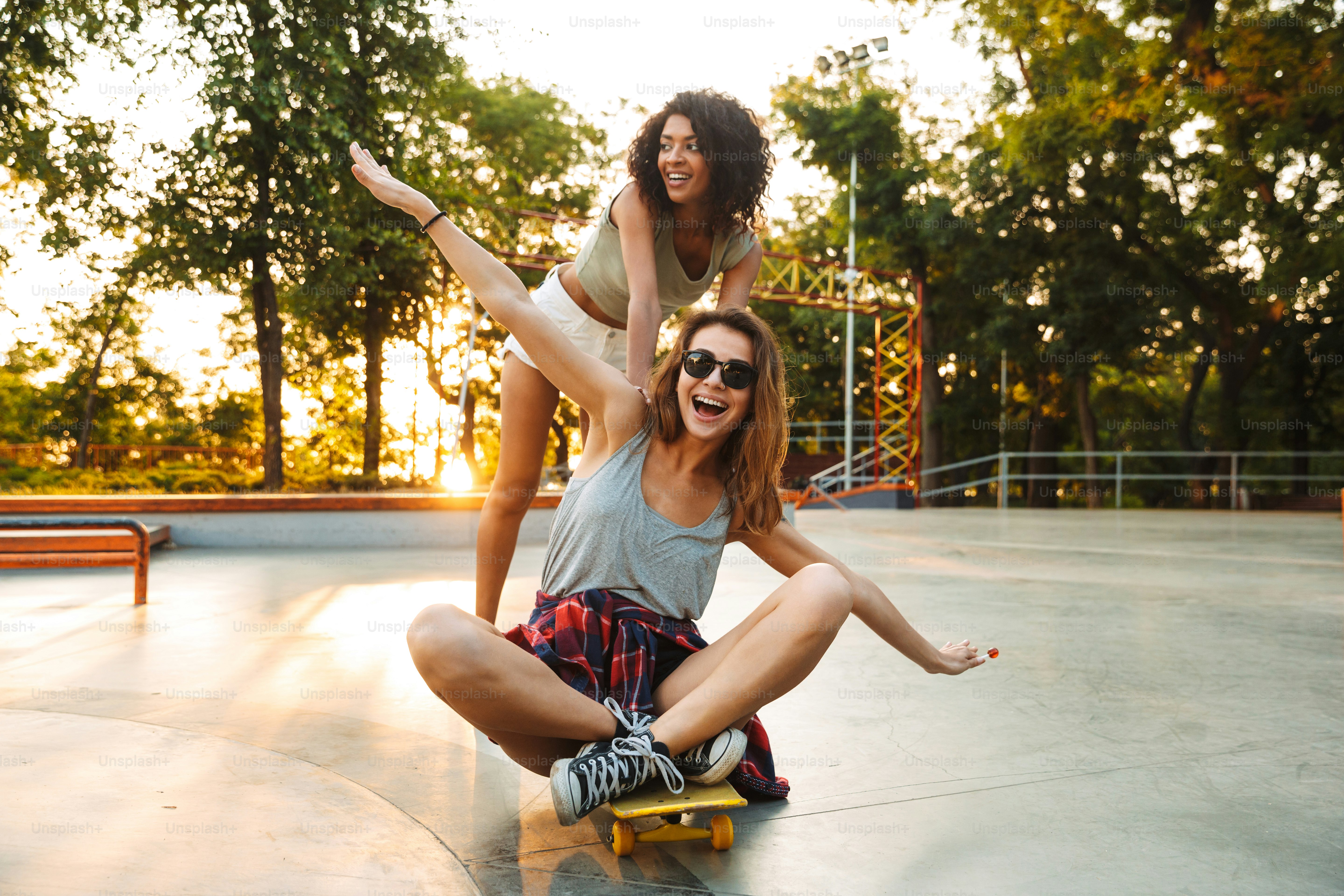 Two positive young girls having fun while riding on a skateboard at the ...