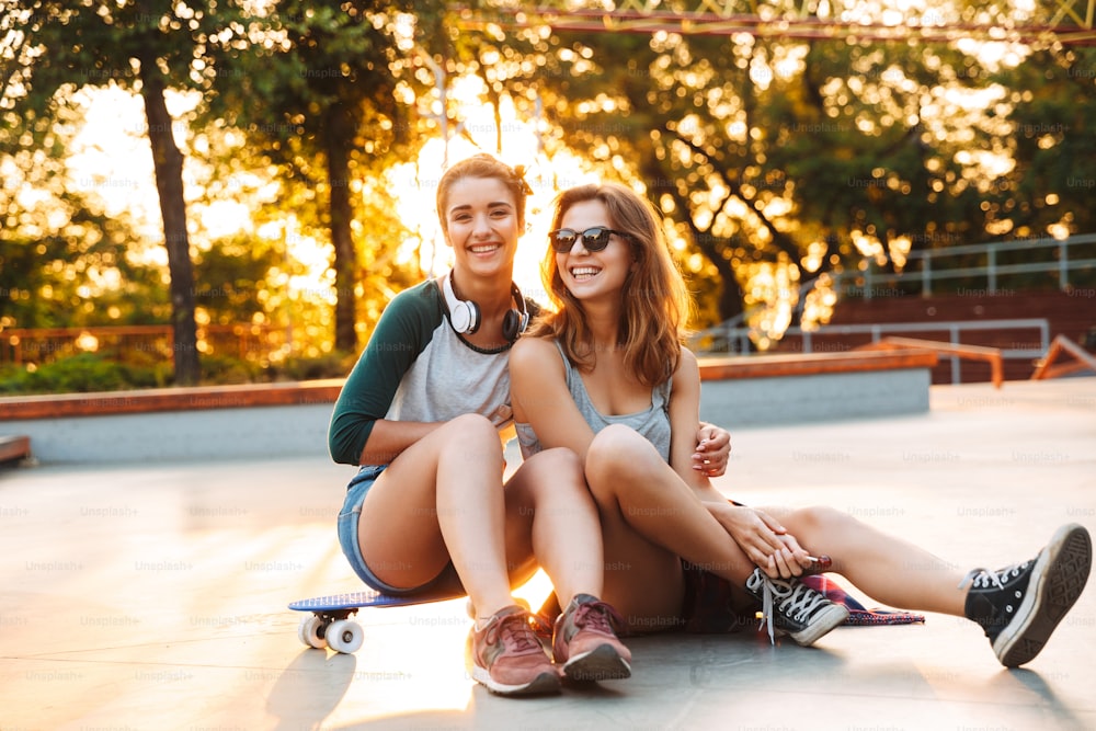 Two smiling young girls having fun while sitting with skateboard at the park