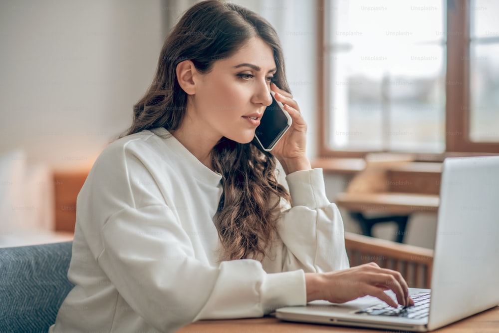 Business woman. Good-looking young woman in a white blouse working on a laptop and talking on the phone